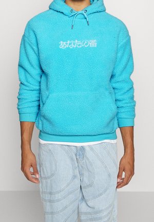 Homme portant un sweat à capuche en polaire turquoise vif avec un texte japonais blanc centré sur la poitrine et un jean bleu clair à motifs.