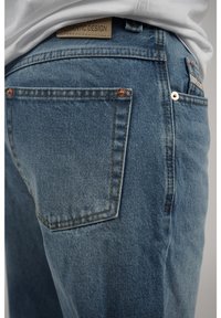 Vaaleansiniset farkut suoran istuvuuden kanssa. Ominaisuuksiin kuuluu oikea takatasku ja kuparinväriset varusteet. Tagi lukee "AITOIN SUUNNITTELU."