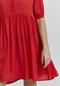 Robe rouge à manches courtes en tissu texturé avec des détails en boutons sur le devant. Jupe évasée avec un ourlet fluide.