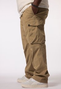 Khaki cargo pants hebben een ontspannen pasvorm, zijzakken en een gladde, duurzame stof; gecombineerd met lichtgrijze sportschoenen.