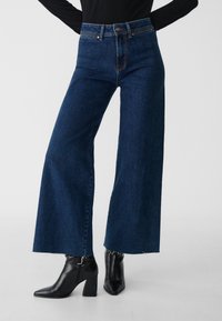 Wide-leg jeans made of dark blue denim with a high waist, featuring a raw hem and contrasting stitching. Paired with black heeled ankle boots.