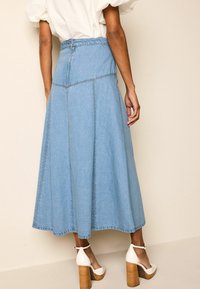 Jupe midi en denim bleu clair avec un design en panneaux et des coutures visibles. Elle présente une fermeture éclair au dos et une coupe évasée. Portée avec des talons blancs.