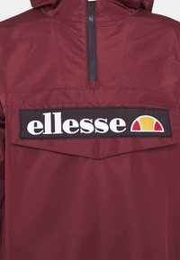 Veste marron avec un écusson noir comportant un texte blanc "ellesse" et un logo en demi-cercle rouge et jaune sous une courte fermeture éclair noire.