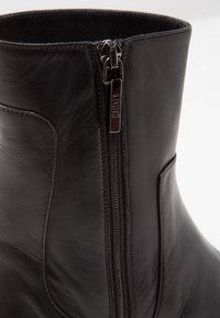 Gros plan sur une botte noire en cuir montant à la cheville, avec une fermeture éclair verticale sur le côté, dotée d'un tirette métallique.