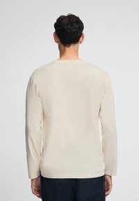 Camicia a maniche lunghe di colore beige chiaro, con colletto rotondo e taglio dritto, caratterizzata da un tessuto morbido e dettagli di cucitura minimi sul retro.
