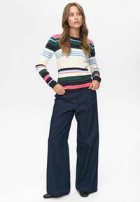Prugasta pletena svetrovka v krem, mornarsko modri, zeleni, rožnati in modri; usklajena s ohlapnimi temnimi kavbojkami z vidnimi šivi.