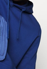 Felpa blu con coulisse, dotata di una tasca testurizzata di una tonalità di blu più chiara. Il materiale sembra morbido e liscio.