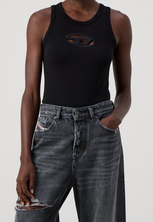 Person wearing a sleeveless black top with oval cutout and dark high-waisted ripped jeans, hand in pocket on light background.