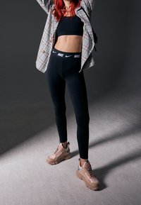 Osoba s červenými vlasy ve kostkované bundě, černém crop topu, černých legínách Nike a robustních béžových teniskách stojící na šedé podlaze.