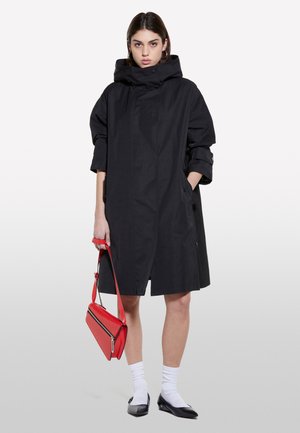 Manteau noir oversize avec capuche, fermeture à boutons sur le devant et poches latérales. Le modèle tient un sac à main rouge. Des chaussettes blanches et des ballerines noires complètent le look.