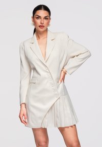 Femme aux cheveux foncés portant une robe-blazer blanche asymétrique avec une jupe plissée, des boucles d'oreilles dorées et des bracelets, posant avec la main sur la hanche.