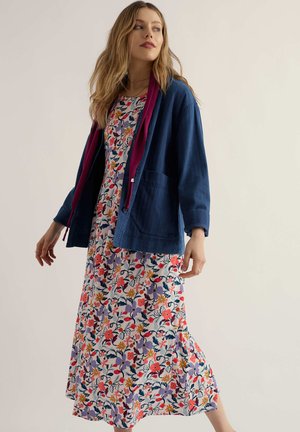 Žena v kvetinových maxi šatách s fialovými, červenými a oranžovými kvetmi, v kombinácii s tmavomodrou bundou s bordovým lemovaním, stojaca proti jednému pozadiu.