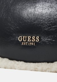 Borsa in ecopelle blu navy con bordo in pelliccia color crema e logo in tono oro "GUESS EST 1981". Texture liscia e forma curva.