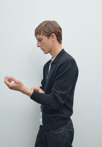 Junger Mann in dunkler, lässiger Jacke und Jeans, der sein linkes Handgelenk an einem schlichten, hellen Hintergrund anpasst, im Profil nach links gerichtet.