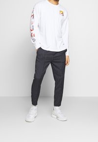 Vit långärmad t-shirt med färgglada mönster på ärmarna, kombinerad med svarta randiga byxor och vita sneakers.