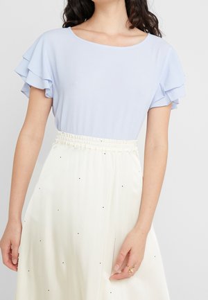 Mujer que lleva una blusa de manga corta azul claro con mangas con volantes y una falda blanca de talle alto con pequeños puntos negros.