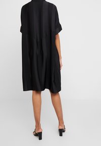 En person klädd i en svart, löst sittande klänning med subtila vertikala ränder, kombinerad med svarta öppna klacksandaler, stående mot en enhetlig bakgrund.