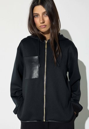 Mujer joven con una sudadera negra con cremallera y un bolsillo de cuero en el pecho, con las manos en los bolsillos laterales, sobre un fondo claro y liso.