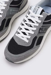 Zapatillas en gris y negro que combinan materiales de malla y ante, con cordones blancos y una etiqueta de logo en la lengüeta. Puntera redondeada y suela acolchada.