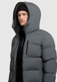 Grijze pufferjack met een grote capuchon. Kenmerken zijn onder andere een ritsborstzak, soepel materiaal en een licht glanzende afwerking.