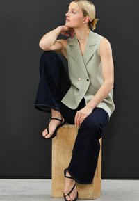 Světle olivový bezrukávový blazer s dvojitým zapínáním a zlatým knoflíkem, spárovaný s tmavými zvonovými džínami a černými sandály na řemínky.