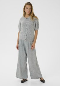 Cardigan en maille grise avec des manches courtes bouffantes et des boutons noirs, associé à un pantalon large à carreaux léger et des chaussures noires plates.