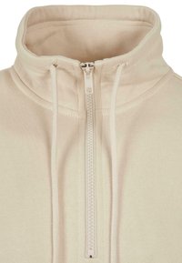 Beige zip-up hettegenser med høy krage, laget av teksturert stoff, med justerbare snorstrammere og frontglidelås.