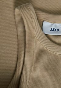 Gros plan sur un débardeur en tissu côtelé beige superposé à un T-shirt, montrant les coutures de l'encolure et une étiquette blanche avec le texte "JJXX".