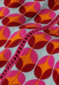 Textil colorido con un patrón geométrico de círculos en rosa, rojo, naranja y azul, con un detalle de puntada texturizada a lo largo del borde.
