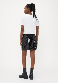 Balta apkirpta marškiniai derinami su juodu blizgiu mini sijonu. Modelis dėvi juodas aulinukus, kurie demonstruoja lygų, blizgantį paviršių.