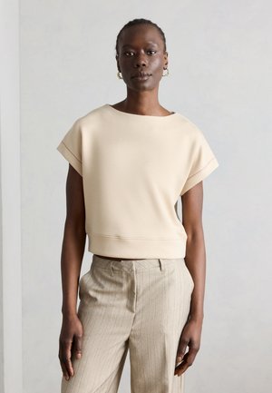 Dunkelhäutige Person trägt ein beiges Kurzarmoberteil und beige Nadelstreifenhosen und steht vor einem schlichten hellgrauen Hintergrund.