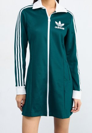 Kobieta ubrana w ciemnozieloną sukienkę zapinaną na zamek marki Adidas z białym kołnierzykiem, białymi paskami na rękawach oraz logo na piersi, stojąca ze swobodnie opuszczonymi rękami.