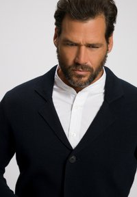 Blazer de punto azul marino con un cierre de un solo botón, solapas con muesca, llevado sobre una camisa blanca de cuello, tejido texturizado, diseño ajustado.