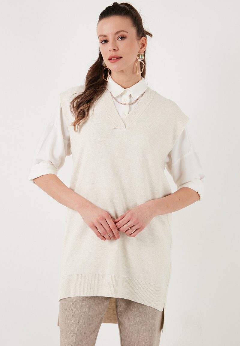 Beige ærmeløs strikbluse med V-hals og ribbede kanter, lagt over en hvid skjorte med krave. Glat tekstur; uformelt design.