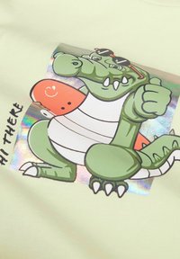 Camiseta de color verde claro con un cocodrilo de dibujos animados que lleva gafas de sol y una patineta. Detalles holográficos rodean la gráfica.