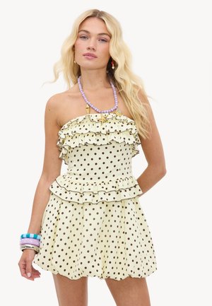 Femme blonde portant une robe jaune à pois sans bretelles avec des volants, un collier de coquillages perlés et des bracelets colorés à son poignet gauche.