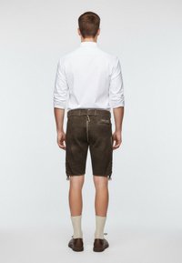 Braune Lederhosen mit schmalem Schnitt, Seitentaschen und dekorativen Bändern am Saum, kombiniert mit einem weißen Langarmshirt und hellen Socken.