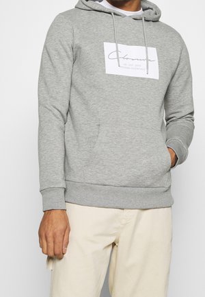 Mężczyzna ubrany w jasnoszarą bluzę z kapturem z przednią kieszenią i białym, prostokątnym logo z napisem „Clozone UK EST 2013 MODERN ESSENTIALS”, w połączeniu z beżowymi spodniami.
