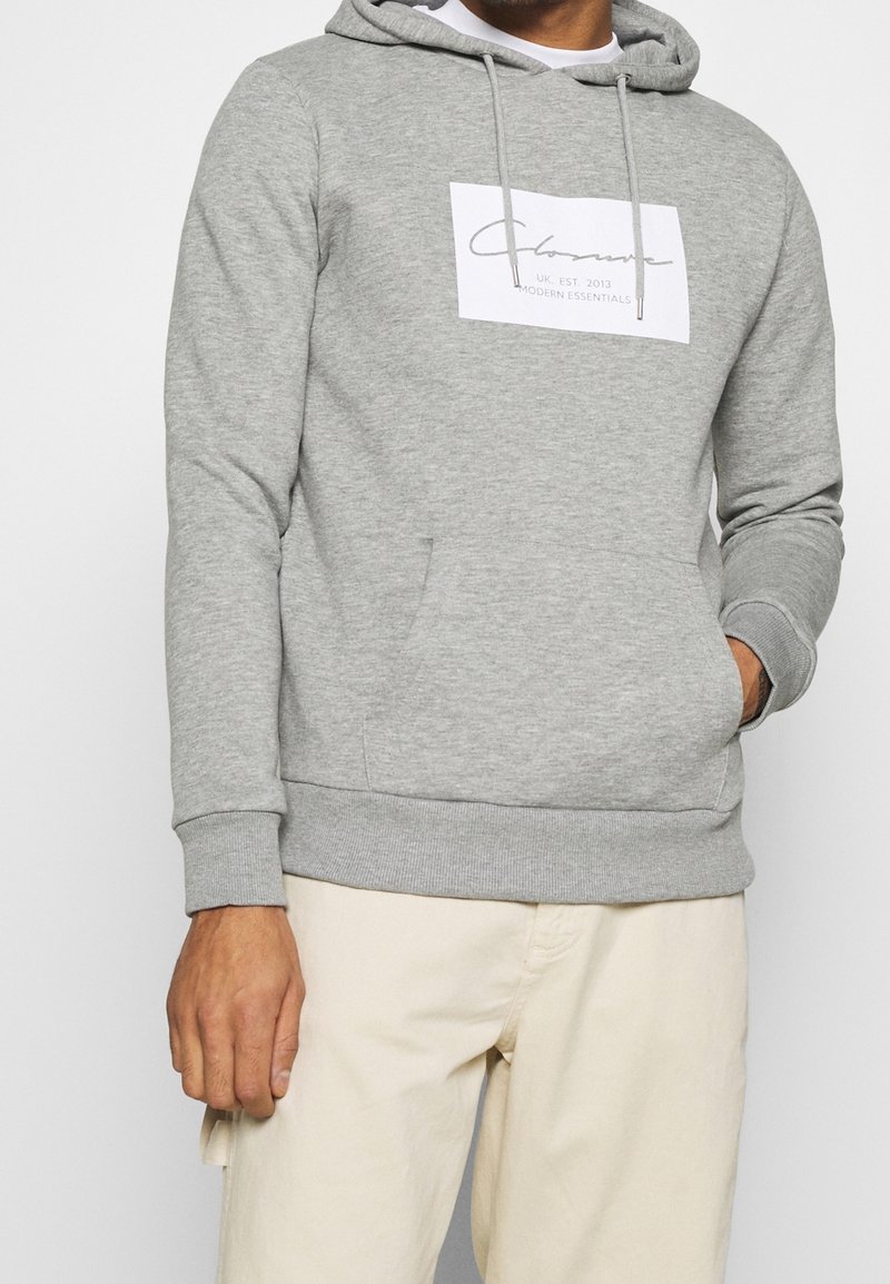 Homme portant un sweat à capuche gris clair avec une poche avant et un logo rectangulaire blanc indiquant « Clozone UK EST 2013 MODERN ESSENTIALS », associé à un pantalon beige.