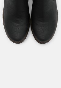 Botas de tobillo de cuero negro con acabado texturizado, puntera redondeada y borde en contraste marrón a lo largo de la suela. Diseño simple y funcional.
