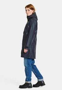 Mörk vattentät regnrock med huva, långa ärmar och justerbara ärmslut, bärs över blå jeans och svarta ankelstövlar, mot en vit bakgrund.