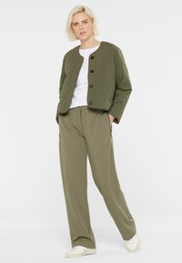 Frau mit kurzem blonden Haar, trägt eine gesteppte olivgrüne Jacke, ein weißes T-Shirt, lockere olivfarbene Hosen und weiße Sneaker, steht mit den Händen in den Taschen.