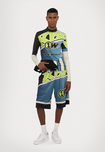 Ensemble de sport multicolore comprenant un haut à manches longues avec des motifs géométriques verts et noirs, associé à un short bleu avec des logos et des accents.
