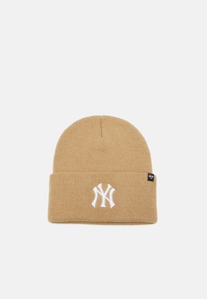 Gorro de punto beige con puño doblado, que presenta un logotipo bordado en blanco en la parte delantera y una etiqueta de marca negra en el lateral.
