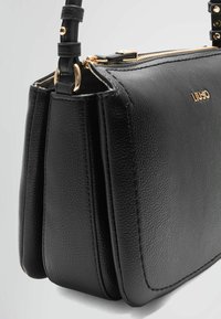 Sac à main en cuir noir avec une finition texturée, doté d'une fermeture éclair dorée et d'un logo. Le design présente une forme structurée avec une bandoulière amovible.