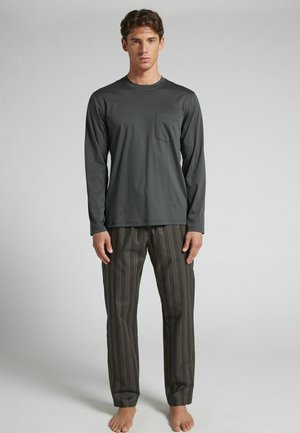 Junger Mann steht barfuß, trägt ein dunkles Langarmshirt mit Tasche und gestreifte, weit geschnittene Hosen vor einfachem Hintergrund.