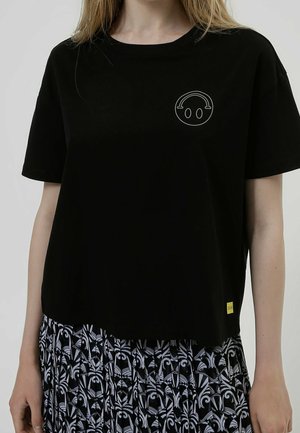 Femme portant un t-shirt noir avec un petit visage souriant blanc et une jupe à motif noir et blanc.