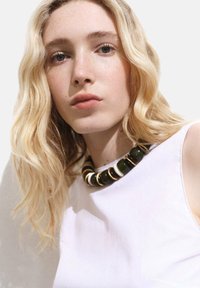 Necklace s crno-bijelim uzorkom sastavljen od velikih perli, s sjajnim završetkom. Model nosi bijelu majicu, izrađenu od mekog, glatkog materijala.