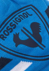 Modrá pletená látka obsahuje černé logo Rossignol s siluetou kohouta a kruhovým okrajem, doplněné trojúhelníkovými bílými a černými vzory.