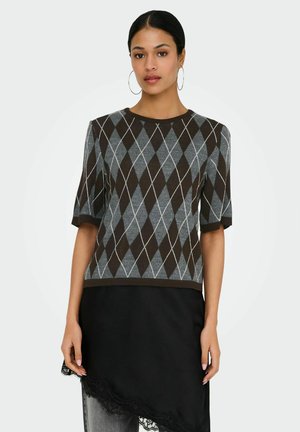 Femme portant un pull à manches courtes à motif losanges marron et gris, et une jupe asymétrique noire avec bordure en dentelle.